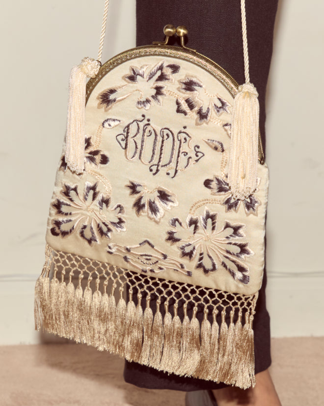 The Phantasm Fringe Bag features ivory silk satin, Victorian-inspired floral embroidery, a "BODE" monogram, tassel details, and a hanging strap.