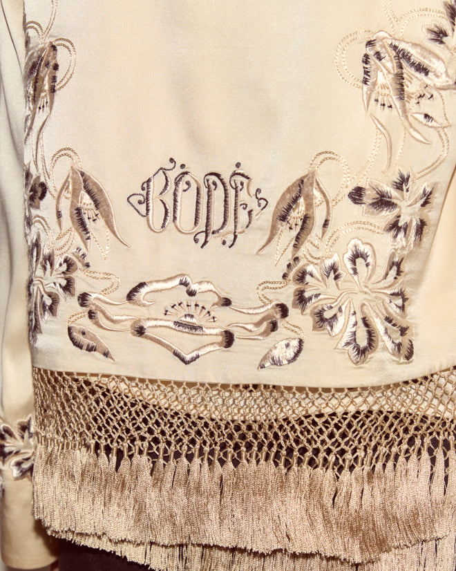 The Phantasm Fringe Jacket features beige embroidered fabric with floral patterns, fringe accents, and "GOD" stitched in the center, evoking the style of Victorian embroidery.