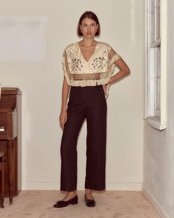 A woman stands indoors by a window and piano, wearing black pants, a cream embroidered top, and draped in the ivory Phantasm Fringe Shawl featuring delicate Victorian embroidery.