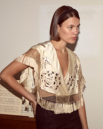 Wearing the Phantasm Fringe Shawl over a cream fringed top and black skirt, a woman stands by sheet music, gazing to the side.