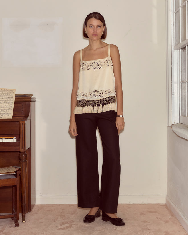 Woman wears the Phantasm Fringe Tank—an ivory silk satin, floral-patterned top—with black pants, standing indoors by a piano and window.