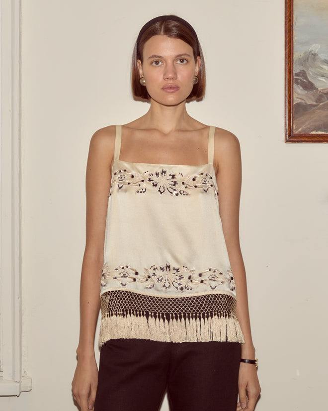 Woman wearing the Phantasm Fringe Tank, a cream top with Victorian-inspired embroidery and tassel details, standing indoors near a painting.