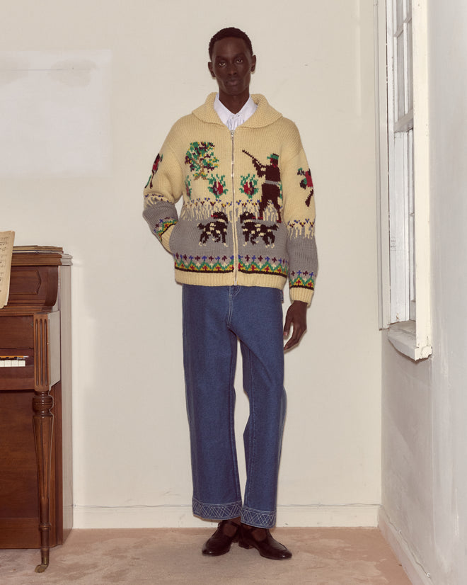 A person stands indoors wearing wide-leg jeans and a Pheasant Cardigan, a cotton knitwear sweater featuring animal and tree designs.