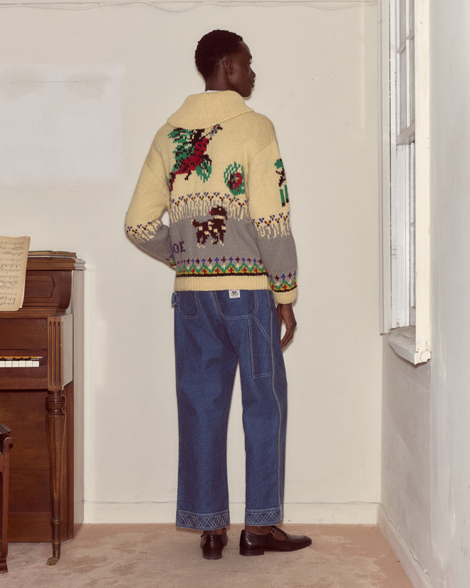 A person stands indoors, facing away, wearing wide-leg jeans and the Pheasant Cardigan, a 1960s-style patterned cotton knitwear piece, near a piano.