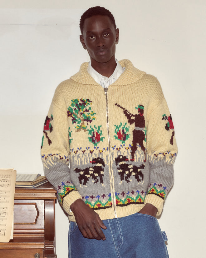 Person wearing a Pheasant Cardigan with a 1960s-inspired pattern stands indoors by a piano with sheet music, looking at the camera.