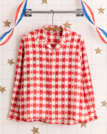 The Picnic Party Shirt, a red and white checkered button-up with a classic plaid pattern, hangs on a rack with festive star and streamer decorations in the background.