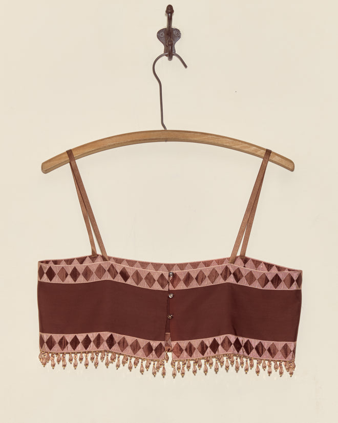 The Pierrot Bead Bra - S, a beaded brown crop top with thin straps made from deadstock ribbon bra material, hangs on a wooden hanger against a plain wall.