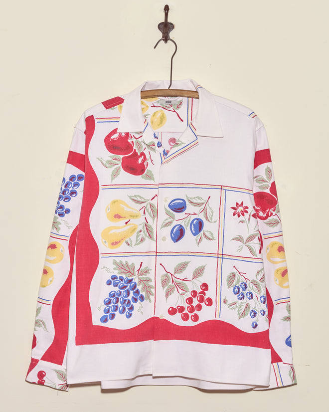 The Pineapple Glory Shirt - L/XL, featuring a vibrant fruit pattern, hangs on a hook against a plain wall.