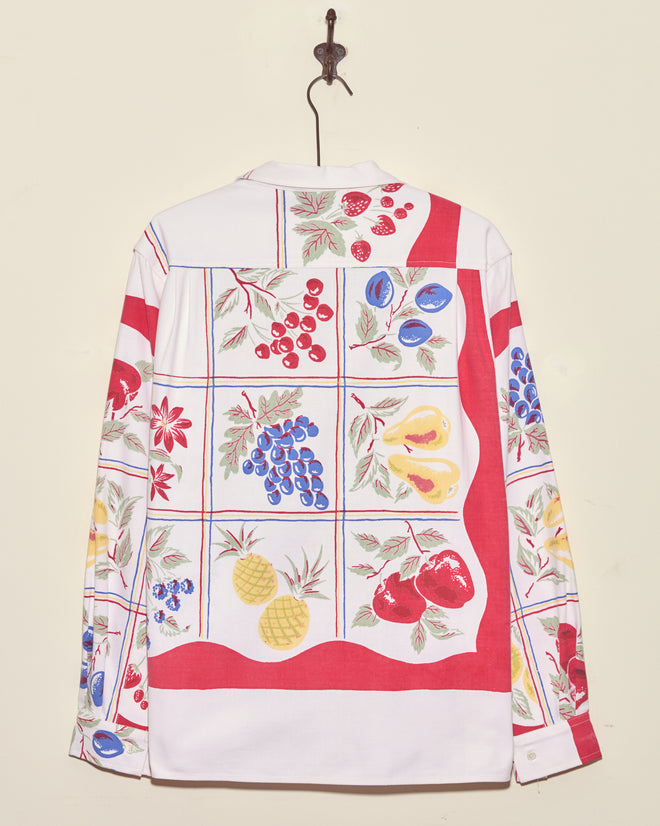 The Pineapple Glory Shirt - L/XL is a unique white long-sleeve with colorful fruit patterns and red borders, reminiscent of a vintage tablecloth shirt, displayed elegantly on a wall hook.