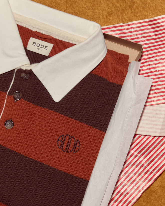 The Pine Stripe Polo, crafted from virgin wool, is presented in a box with tissue paper and a red striped bag on a brown surface.