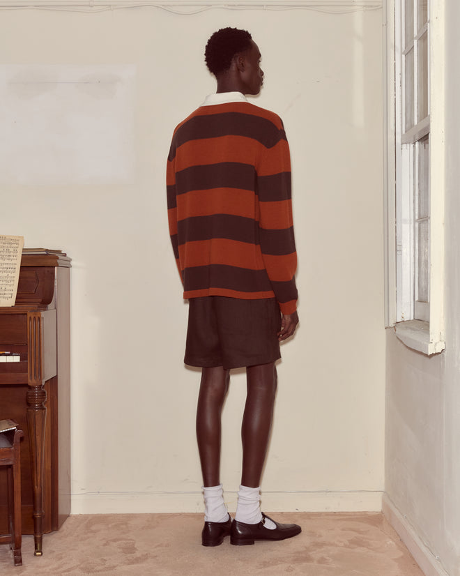 A person in a Pine Stripe Polo and shorts stands facing a wall in a room with a piano and a window.