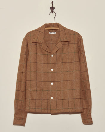 The Pinscher Check Overshirt - M/L is a unique, brown plaid button-up with a collar, crafted from deadstock wool and displayed hanging on a hook against a plain, light wall.