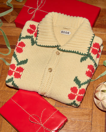 A folded cream Pirate Ship Cardigan with red floral patterns sits among wrapped red gifts on a wooden floor.