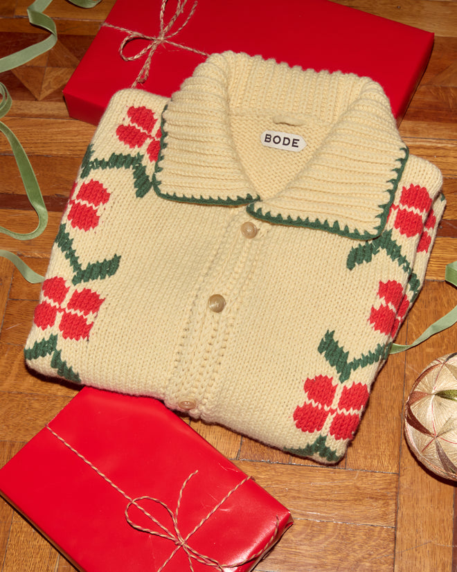 A folded cream Pirate Ship Cardigan with red floral patterns sits among wrapped red gifts on a wooden floor.