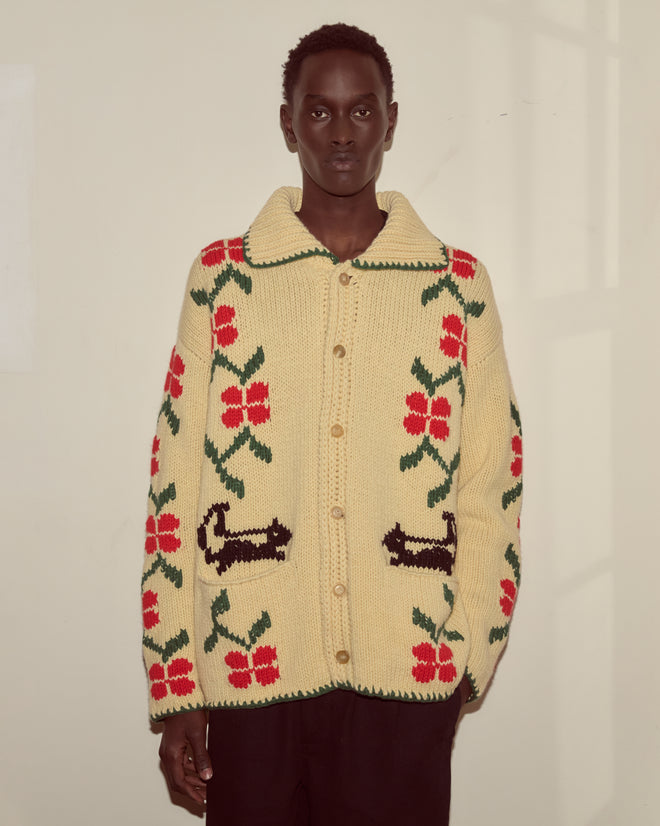 A person wears the Pirate Ship Cardigan, a cream sweater featuring red flowers, black animal patterns, and a playful pirate ship motif, standing against a beige wall.
