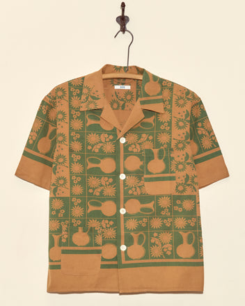 The Pitcher Print Short Sleeve Shirt (XS/S) features a unique green and tan floral pitcher pattern and is displayed hanging on a decorative wall hook.
