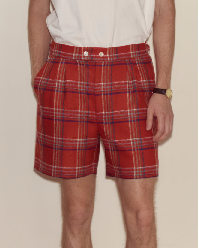 A person wearing Gable Plaid Shorts in red, a white shirt, and a watch, with hands in pockets.