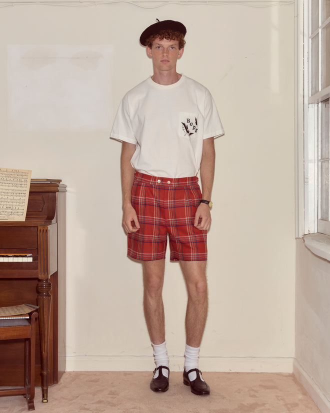 A young man in a beret, white t-shirt, Gable Plaid Shorts, and dress shoes stands by a piano indoors.