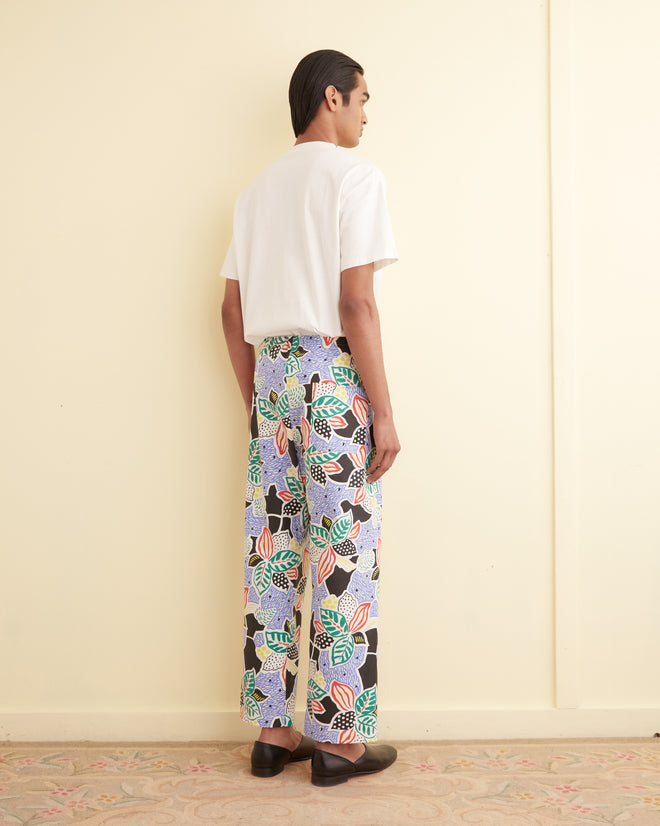 A person stands facing a cream wall, wearing a white shirt and colorful Plantain Jazz Pajama Pants.