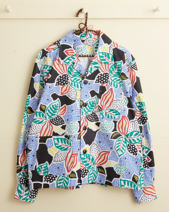 The Plantain Jazz Long Sleeve Shirt is a unique button-up with bold, oversized botanical and abstract patterns, shown hanging on a hook against a wall.