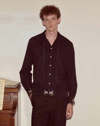 A man wearing the Pointer Long Sleeve Shirt in black, featuring subtle ladderwork details, stands by a piano with sheet music against a plain wall.