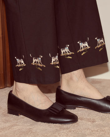 Person wearing Pointer Side-Buckle Trousers—straight leg black chinos with an embroidered dog motif near the cuffs—paired with black loafers featuring a bow detail.