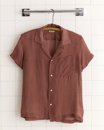 The Polo Dot Shirt is a short-sleeve, brown button-up featuring a unique white dot pattern, displayed on a hanger against a white tiled wall.