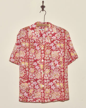 Pomegranate Leaf Short Sleeve Shirt (M/L) featuring a yellow and white floral pattern on red fabric, short sleeves, and button-up front, displayed hanging on a hook against a wall.