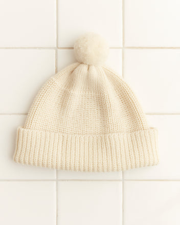 The Pom Hat - Cream, a vintage-style knit beanie with a pom-pom on top, is crafted from soft merino wool and elegantly placed on a tiled white surface.