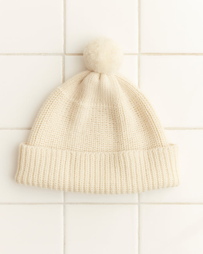 A Pom Hat - Cream with a vintage knit design and a cream pom-pom rests on white tile, showcasing its classic charm.