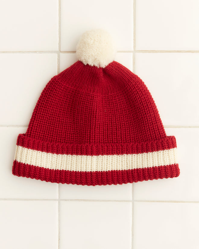 The Pom Hat - Red showcases a charming vintage knit design in red, accented with a cream stripe and a playful pom-pom, all set against a tiled background. Crafted in Peru, this comfortable accessory is made with premium merino wool for unparalleled style and coziness.