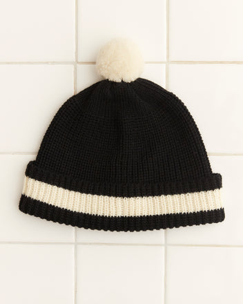 The Pom Hat - Black is a vintage knit cap showcasing a black design accented with a white stripe and topped with a playful pom-pom, crafted from soft merino wool, and displayed on a tiled surface.