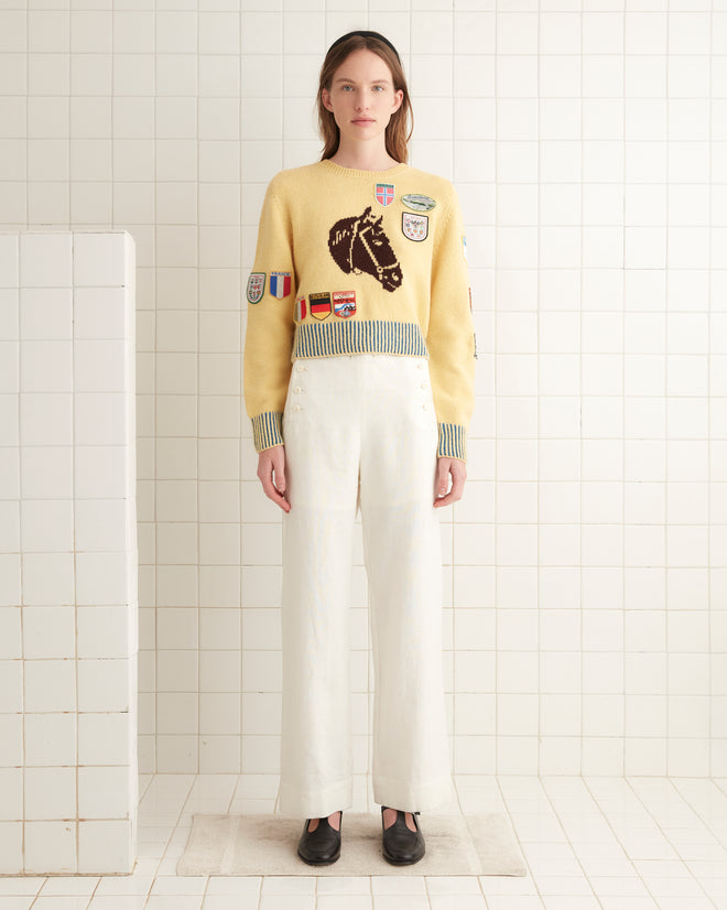 A person wearing a yellow Pony Silhouette Crewneck adorned with vintage patches and white pants stands in a tiled room.