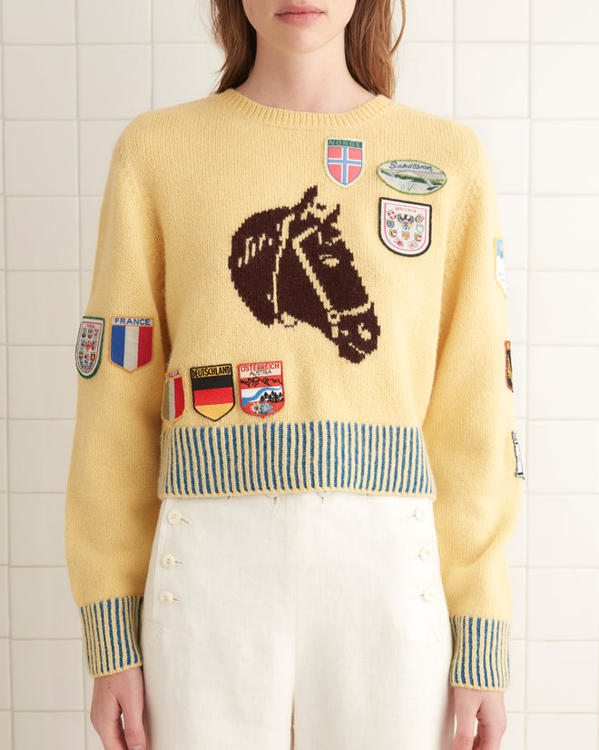 A person wearing a yellow Pony Silhouette Crewneck adorned with vintage patches and white pants stands in a tiled room.