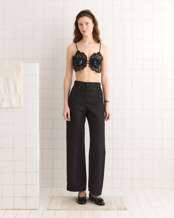 In a tiled room, a woman wears the 1940s-inspired Poppy Bra - Black with circular designs, paired elegantly with high-waisted black pants.