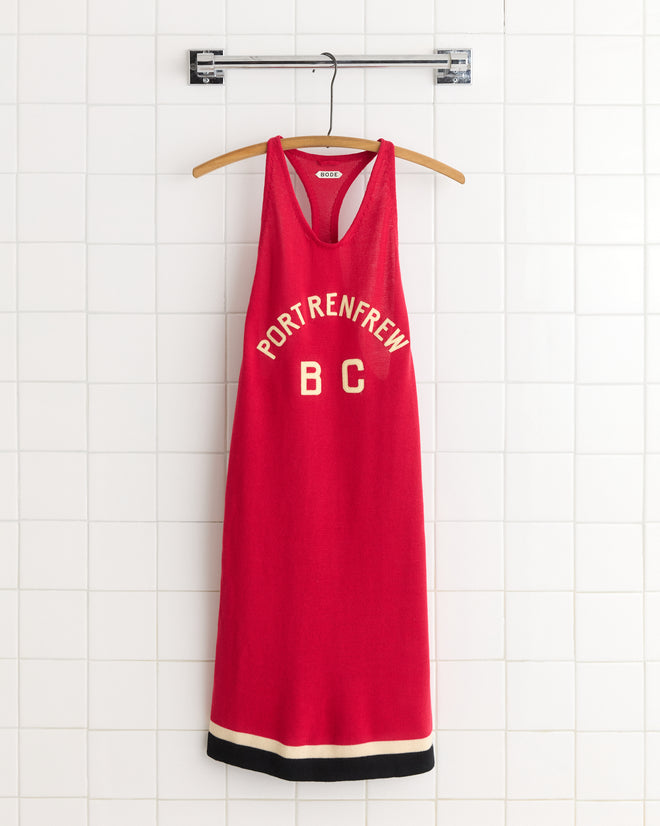 The Port Dress is a red sleeveless piece inspired by 1900s-1920s rowing singlets, featuring "PORT RENFREW BC" text and shown on a wooden hanger against a white tiled wall.