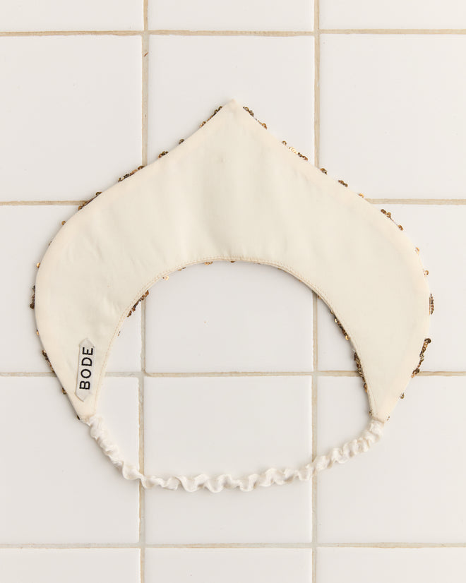 The Pinnacle Crown, a white fabric collar adorned with bronze and silver sequins, labeled "BODE," rests elegantly on a tiled surface.