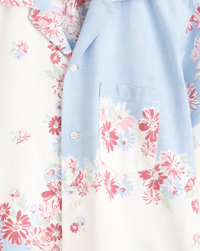 The Powder Blue Floral Shirt - S/M, a short-sleeved, button-up adorned with vibrant floral patterns in shades of pink and white on light blue and white fabric, reminiscent of a midcentury tablecloth, hangs on a hanger against a cream-colored wall.