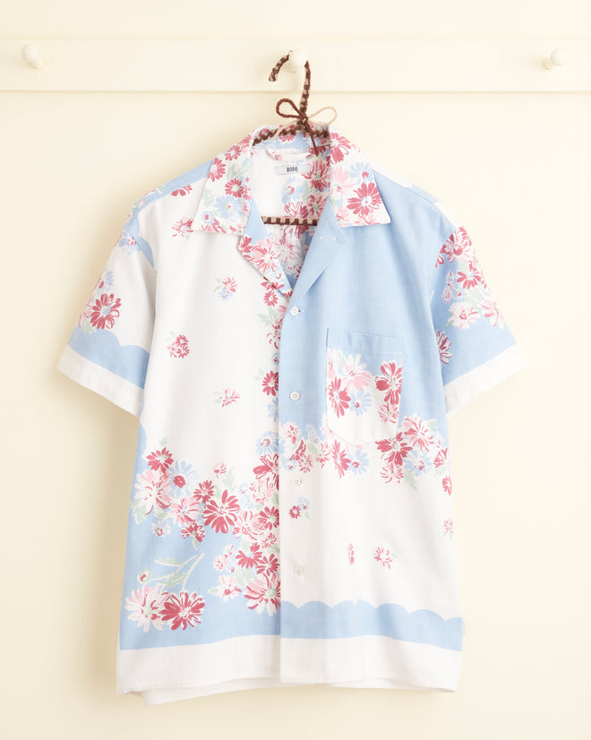 The Powder Blue Floral Shirt - S/M, a short-sleeved, button-up adorned with vibrant floral patterns in shades of pink and white on light blue and white fabric, reminiscent of a midcentury tablecloth, hangs on a hanger against a cream-colored wall.