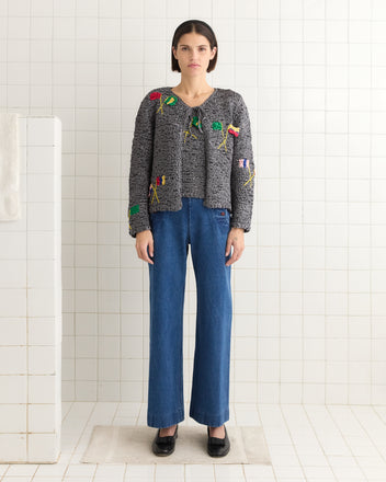 In a tiled room, a person stands confidently, wearing the Promenade Cardigan with classic blue jeans.