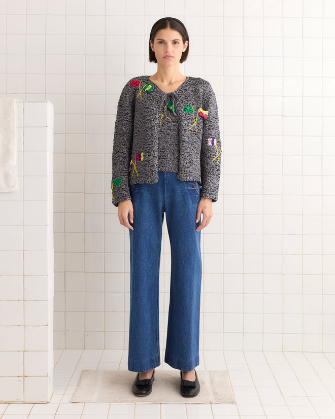 A person wearing a Promenade Cardigan, inspired by 1950s knitwear with colorful textured gray patterns, stands in a tiled room with blue jeans and black shoes.