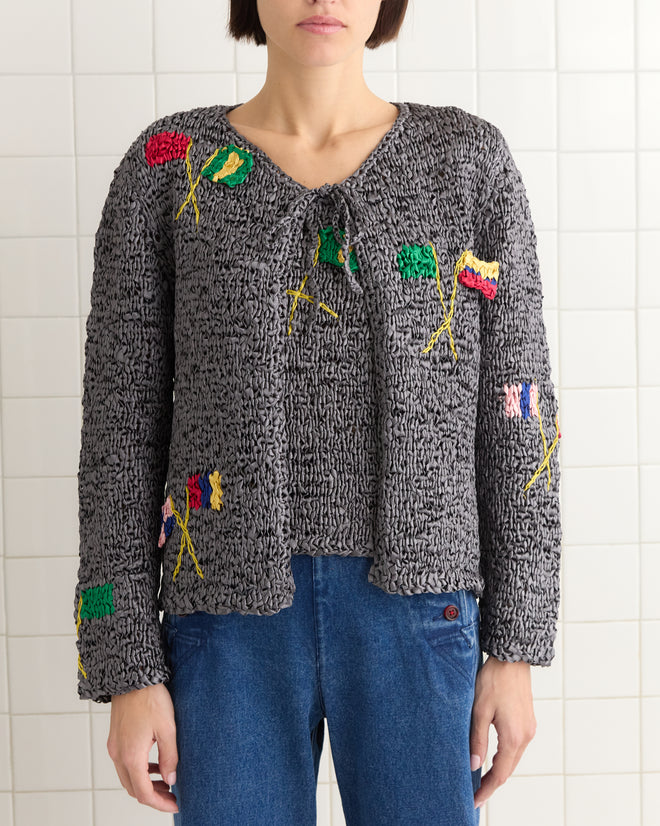 A person wearing the Promenade Cardigan, a gray textured sweater with colorful 1950s-inspired embroidered designs, stands indoors paired with blue jeans.
