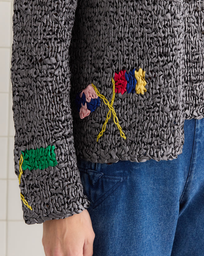Close-up of a person wearing the Promenade Cardigan, featuring textured gray fabric with colorful embroidery inspired by 1950s reproduction styles, elegantly paired with blue jeans.