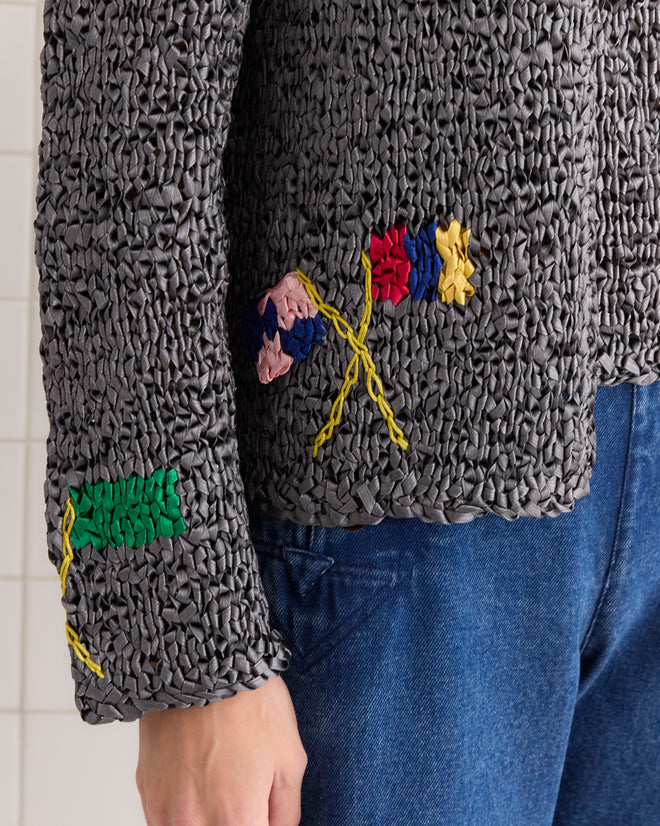 A person wears the Promenade Cardigan, a textured gray knitwear top with colorful abstract embroidery, elegantly paired with blue jeans.