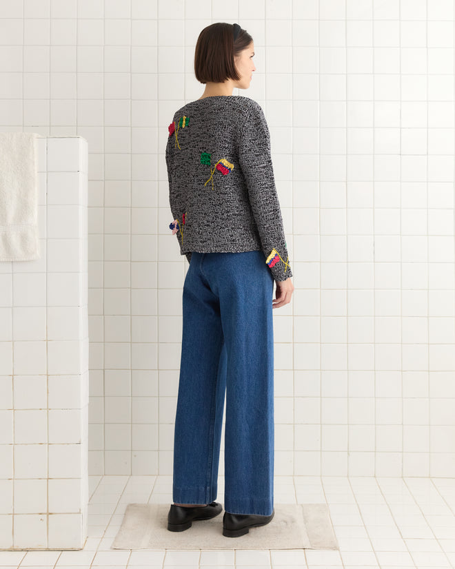 In a tiled room, a person wearing the Promenade Cardigan, featuring colorful 1950s-inspired designs, stands facing away, perfectly matched with blue wide-leg jeans.