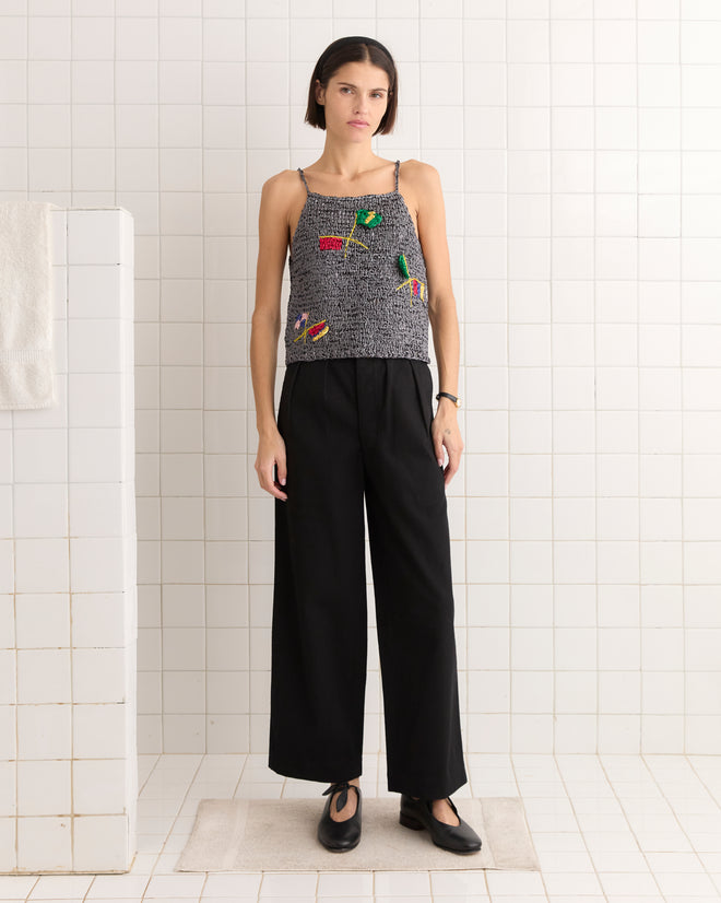 Wearing a Promenade Tank in black and gray that mirrors sportswear chic, the individual stands by a tiled wall, paired with sleek black pants.