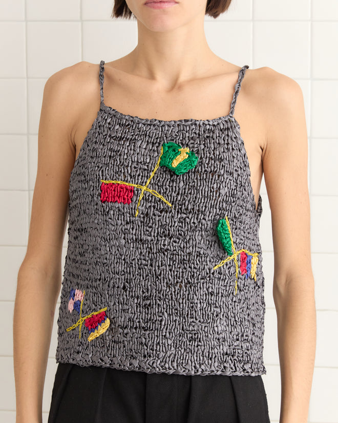 A person is standing against a tiled wall, wearing the Promenade Tank, a textured gray top with colorful abstract embroidery that resembles ribbonwork crochet.