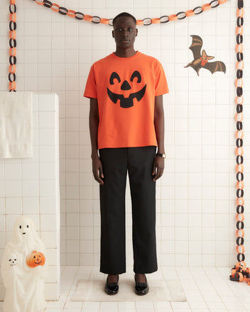 The Pumpkin Tee, a vintage Halloween shirt featuring an iconic jack-o'-lantern pattern and crafted from 100% cotton, hangs on a white wall hook.