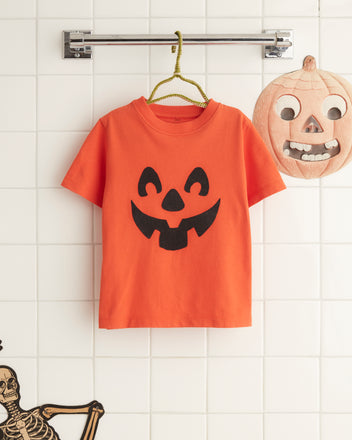 A Pumpkin Kids' Tee featuring a black jack-o'-lantern pattern is displayed on a white tile wall, alongside skeleton and pumpkin decorations.