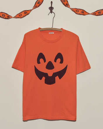 The Pumpkin Tee, an orange shirt with a black jack-o-lantern face, hangs on a hook beneath Halloween garland—ideal for anyone who loves festive Halloween shirts.